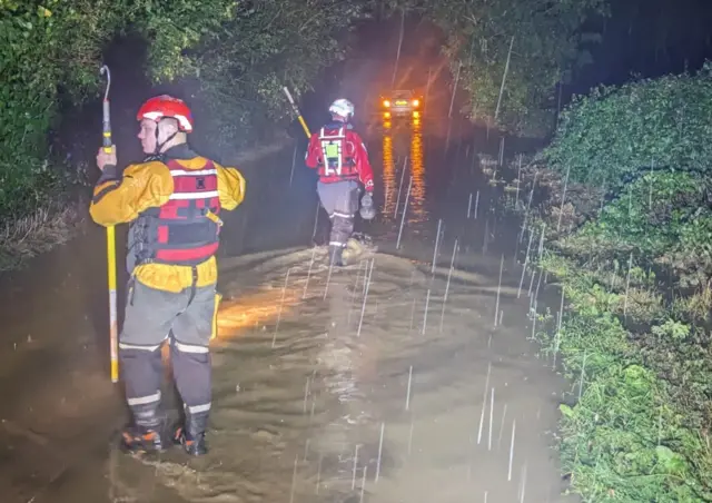 Firefighters are seen wading through a flooded road in Northamptonshire. They are wearing specialist water uniforms with lifejackets and helmets. A stranded car can be seen in the distance with its hazard lights on.