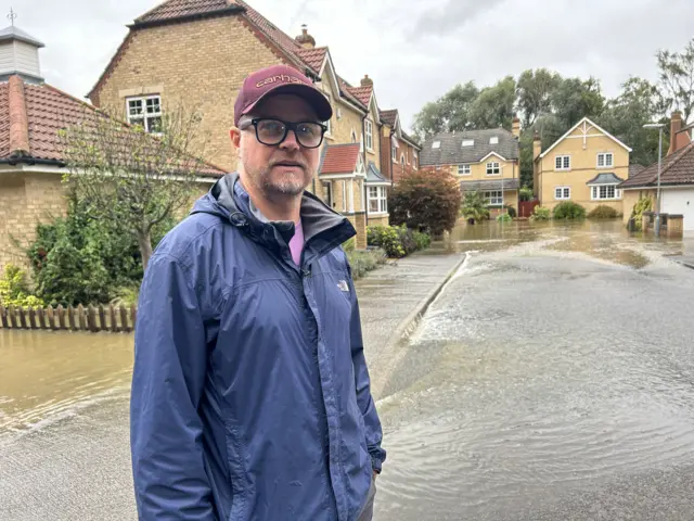 Dean Elcombe in a navy rain coat and a red baseball cap standing in the street.