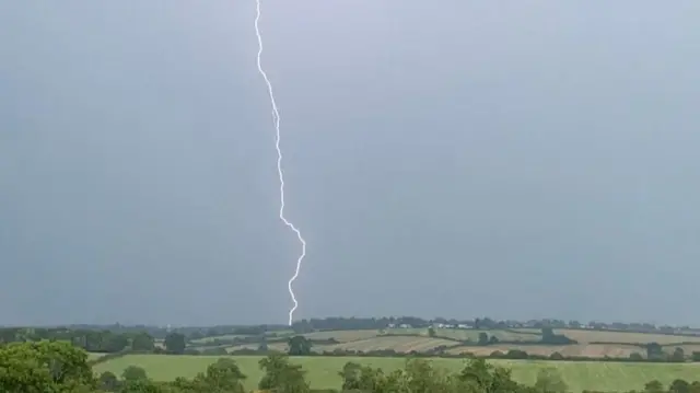 A photo taken from the Northamptonshire countryside shows a single bolt of lightning hitting the ground several miles away. The sky is dark grey with the bolt lighting up the surrounding area. Green, rolling fields can be seen all around with the occasional building.