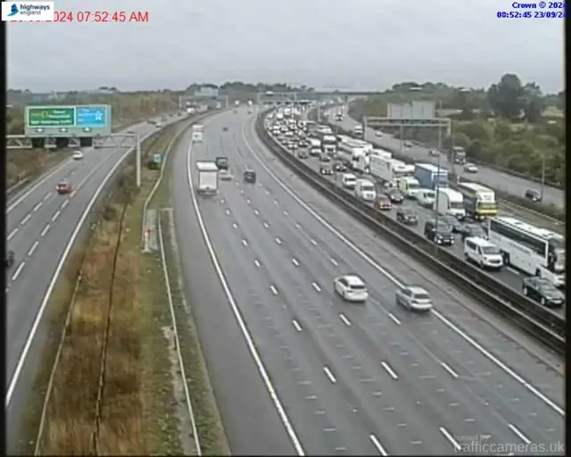 A traffic camera image shows the M1 carriageway. Long queues can be seen on the northbound carriageway. Traffic on the southbound carriageway can be seen moving freely.