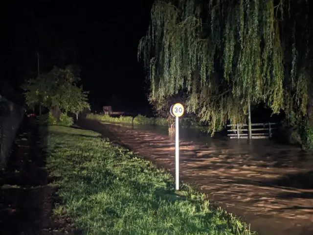 A photo shows a Northamptonshire road late at night that has flooded. Water can be seen covering the entirety of the road while a vehicle's lights shine on the affected area. A road sign can be seen and the grass verged are still visible.