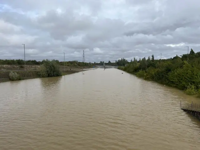 The A421 at the Marston Moretaine Interchange. The picture shows the road completely submerged with water and it looks like a river itself.