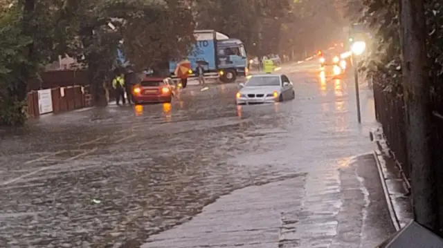 A car makes its way through a flooded street. The water is almost at the top of the tyres. A queue of cars with headlights on can be seen waiting behind it, not entering the big puddle