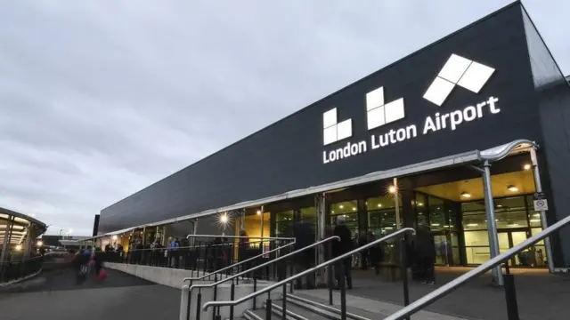 A general view of the exterior and entrance of London Luton Airport. It shows steps leading up to the entrance of the airport. A large sign on the building reads "London Luton Airport"