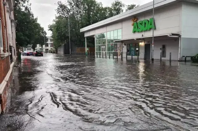 A picture taken from Rushden High Street outside the Asda supermarket. The road can be seen flooded with water outside the supermarket. Cars can be seen in the distance attempting to drive through the water.