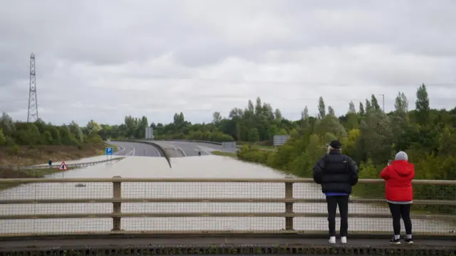 Two people standing on the bridge overlooking the flooded A421 dual carriageway at Marston Moretaine.