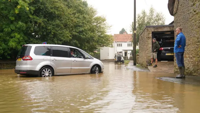 A car driving through brown flood water in Grendon, Northants, with two men standing nearby on