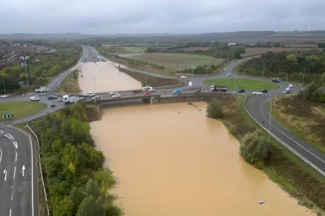 A drone image of the A421 at the Marston Moretaine Interchange in Bedfordshire. The pictures shows the carriageway completely flooded in both directions under a bridge. Cars are driving over the bridge while a single vehicle lies stranded in the flood water beneath.