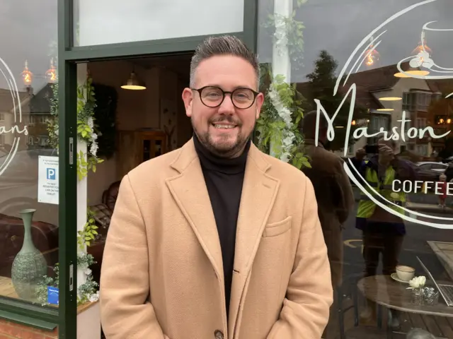 James Kendall is pictured outside a cafe in Marston Moretaine in Bedfordshire. He is smiling to the camera while wearing a light brown coat, black top and black glasses. He has a beard and short brown/grey hair.