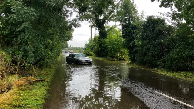 A road is submerged in water, surrounded by trees and bushes either side, with a silver car stuck in the middle