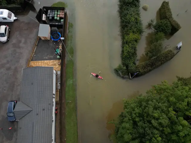 A drone image shows a man canoeing down a flooded street after heavy rain in Northampton. The man can be seen in a red canoe with paddles. Trees and bushes line the road and a house can be seen nearby that has not suffered from any flooding.