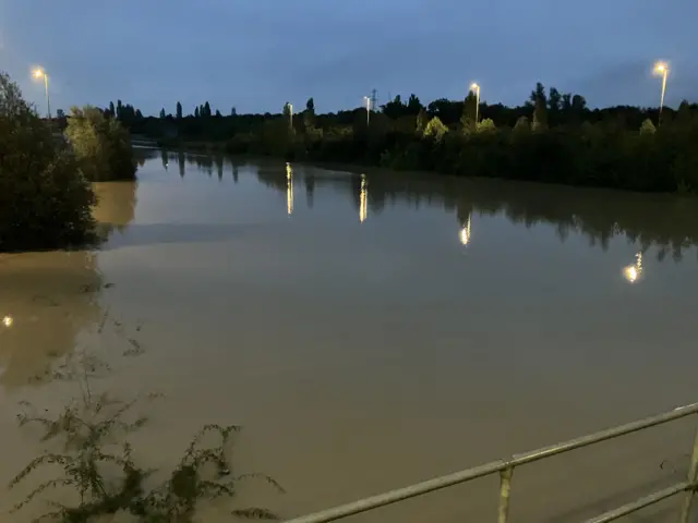 The A421 is pictured during early morning and is completely flooded. It is pictured looking like a river itself and the road's street lamps are reflecting in the water. Trees and bushes line the flooded road.