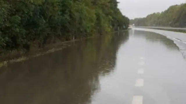 A picture of the flooded A5 carriageway in Buckinghamshire. The picture shows a heavy body of water that has flooded most of the carriageway. The road is lined by trees and no vehicles can be seen on the road.