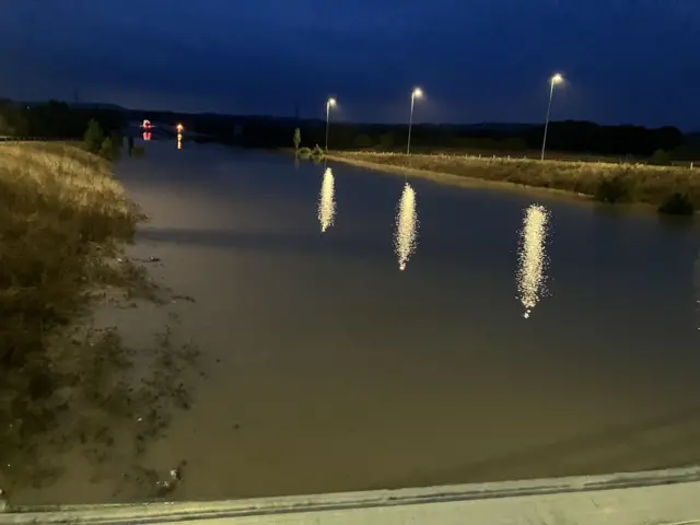 Another angle of the A421 flooded carriageway during early morning when it is still dark. Vehicles can be seen in the distance with their lights on where the road is not flooded. Street lamps reflect in the water while trees and bushes line the flooded road.