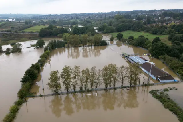 A drone image of Spencer Football Club pitch in Northampton. It shows the pitch and surrounding field that are completely submerged in water. Goal posts and buildings can still be seen.