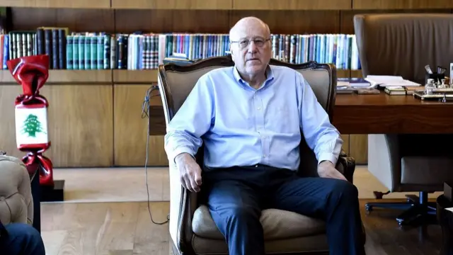 A man in a blue shirt and trousers sits in an armchair with a bookcase behind him