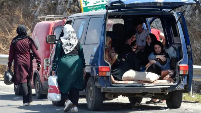 Two women walk beside a van - a group sit inside, with one woman cradling a child