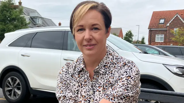 A general view of Rachel Lenton. She is facing and smiling to the camera. She is wearing a leopard-print dress and has brown hair with a blonde streak. She is wearing a watch and her arms are placed one over the other in her lap.