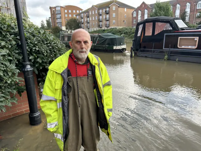 Ian Bates is pictured standing in front of the River Nene in Northampton that has burst its banks. Boats are pictured behind him on the water with blocks of flats in the background. He is wearing green waterproof overalls as well as a high-vis coat.