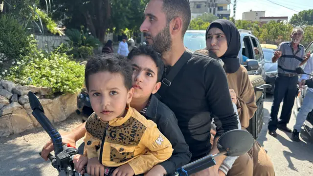 A family of five sit on a motorbike with a green suitcase strapped to the back of it