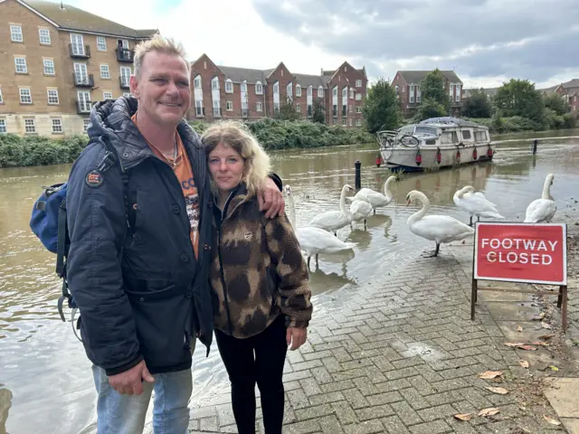 Lee Mills and Saphire Blurton are pictured with their arms around each other in front of the River Nene. The river has burst its banks and there are several swans standing behind them. A sign warning that the footpath is closed is next to them. Several flats are in the background.