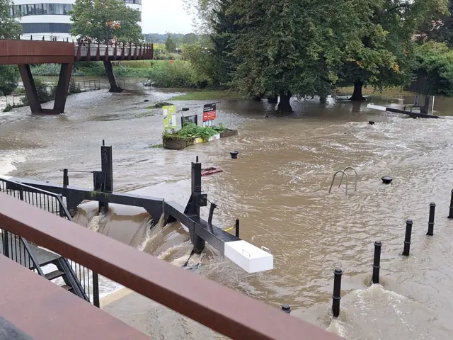 A photo shows the flooding in Northampton after the River Nene burst its banks. Fast flowing water is pictured moving over a lock while grass nearby has been flooded over. An overbridge, a building and trees can be seen in the distance.