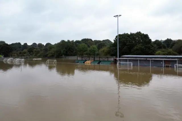 A ground level photo of the Spencer Football Club pitch. The stands and football buildings can be seen as well as goal posts. Trees sit in the background.