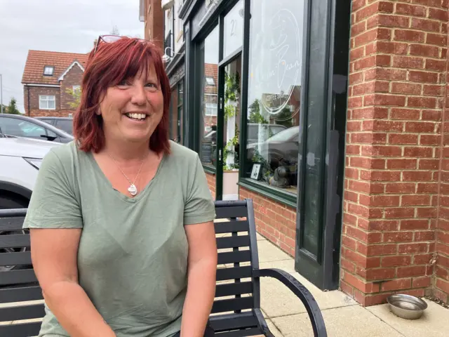 A general view of Kirsty Fanthorpe. She is pictured smiling to the camera and is wearing a green T-shirt with a necklace, red hair and glasses on top of her head. She sits outside a cafe on a bench.