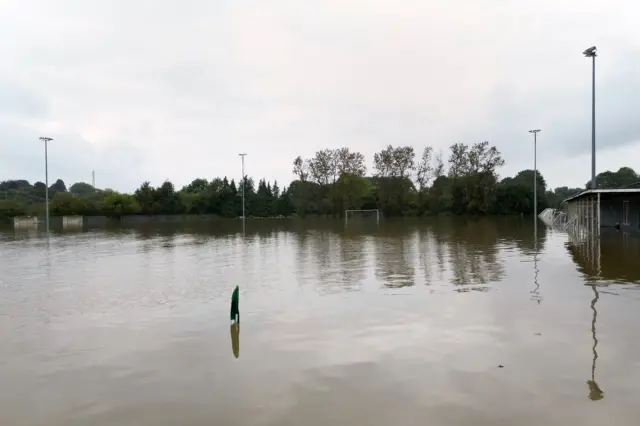 Another view of the flooded Spencer Football Club pitch. The water has completed submerged the area with just the goal posts, pitch lights and stadium buildings on show above the water.