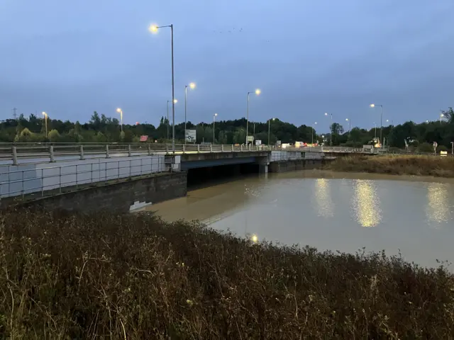 A picture of the A421 road in Bedfordshire taken during the early hours of Tuesday morning. It shows the carriageway flooded under a bridge. Street lamps remain on and they are reflecting in the water. The flooded carriageway is lined by foliage.