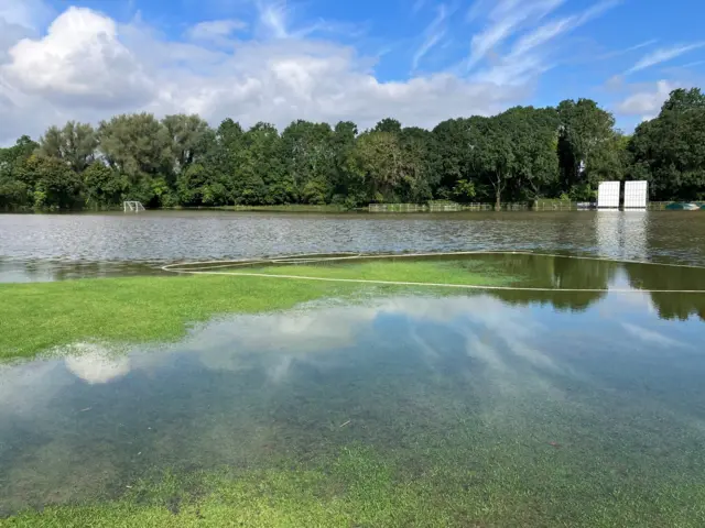 Another angle of the flooded cricket pitch. Parts of the pitch can be seen through the water. Cricket equipment, included white sight screens, is in the background as well as trees that border the pitch.