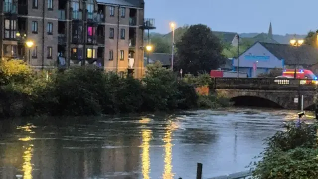 A photo of the River Nene in Northampton. The river's water level is high and a bridge can be seen in the distance with a fire service vehicle on it. Buildings can be seen around the river while lights reflect on the water.