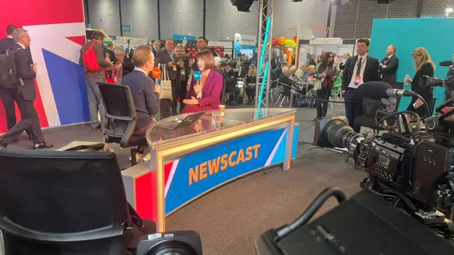 Photo taken behind cameras showing Rachel Reeves being interviewed at a desk reading 'Newscast'