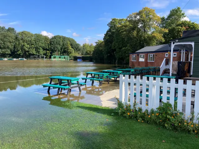 A cricket pitch is pictured under flood water. Picnic benches can be seen in the water along with their feet just covered in water. There is a skatepark and cricket nets in the background on the far side of the pitch.