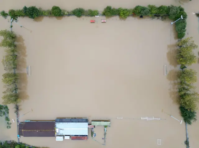 A drone image shows the Spencer Football Club pitch under water in Northampton. The entire pitch is covered with water while the goal posts can be seen at each end of the pitch. The club's buildings can be seen as well as trees and bushes that surround the pitch.