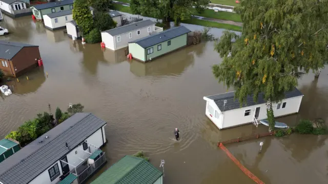 A man is wading through knee-high water in the middle of a holiday park with static homes.