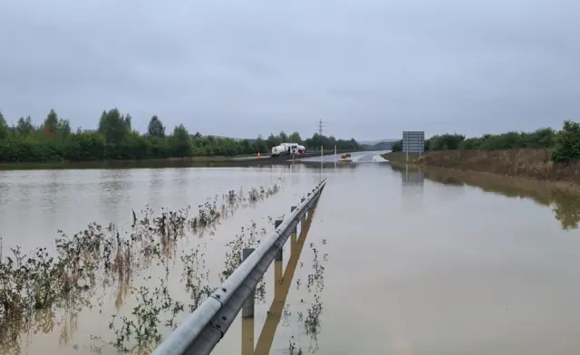 The A421 flooded road in Bedfordshire pictured a day after it was heavily flooded. The road is completely submerged while tankers can be seen in the background that have arrived to pump out water. The non-flooded road ahead has no other vehicles on it. A road sign can be seen in the distance while the road is lined with trees and grass.