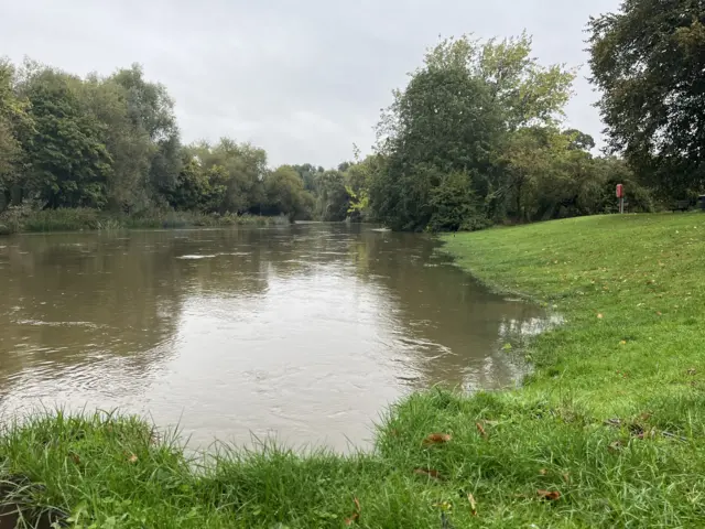 A picture of the Great River Ouse from a different angle that shows it has burst its banks. Water can be seen flooded onto nearby grass while trees line the river further down.