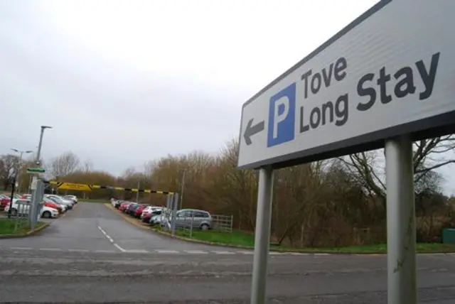 A general view of Tove long stay car park in Towcester. It shows the road sign for the car park with cars parked behind. A barrier can be seen along with street lights.