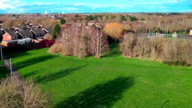 Aerial view of a playing field, surrounded by trees, with houses seen behind the field