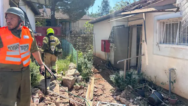 Firefighters in bright jackets stand next to a damaged house with debris at their feet