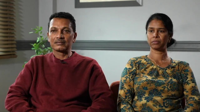 A man in a maroon jumper and a woman in a gold shirt with floral patterns on sit side by side in a room with white walls