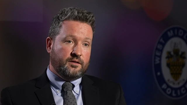 A man in a light blue shirt, grey tie, and black jacket looks towards the camera in front of a background with the West Midlands Police logo