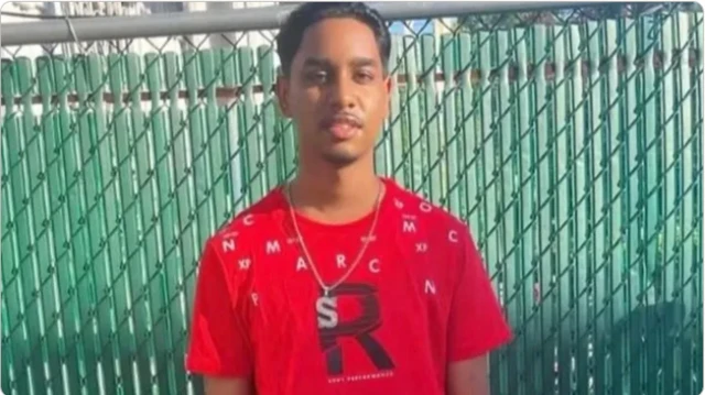 A young man in a red t-shirt with letters printed on it across the shoulders, and a large black r on the chest. The man also wears a silver chain which has a big silver s pendant attached to it. Behind him is a green fence.