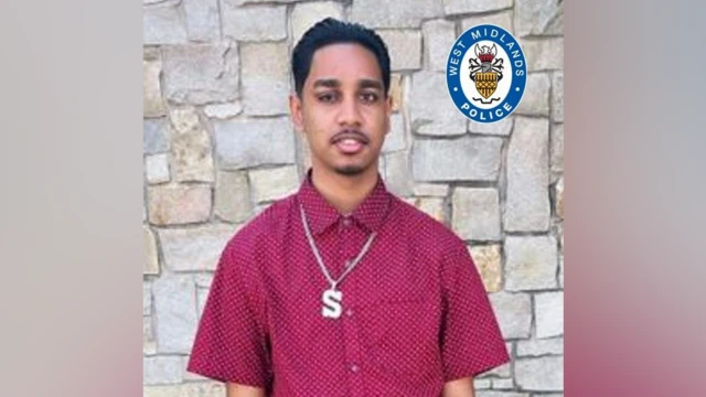 A man in a maroon shirt, wearing a chain with the letter S on it, looks at the camera; he is standing in front of a brick wall