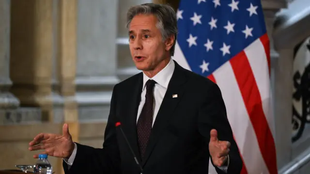 Anthony Blinken standards behind a lectern, wearing a dark suit and with a US flag behind him. He is talking and has his hands raised either side of him