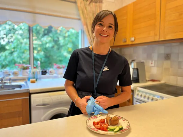 Carer Elizabeth Del Giacco, in a dark t-shirt and ponytail, smiles as she stands at the kitchen counter having prepared Jess's dinner - a pork pie with tomatoes and cucumber, which sits on a plate on the counter