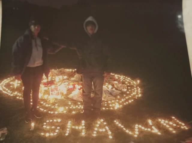 Two people in coats stand on a field in the dark, surrounded by tealights and flowers; tealights spell out the name 'Shawn' on the grass