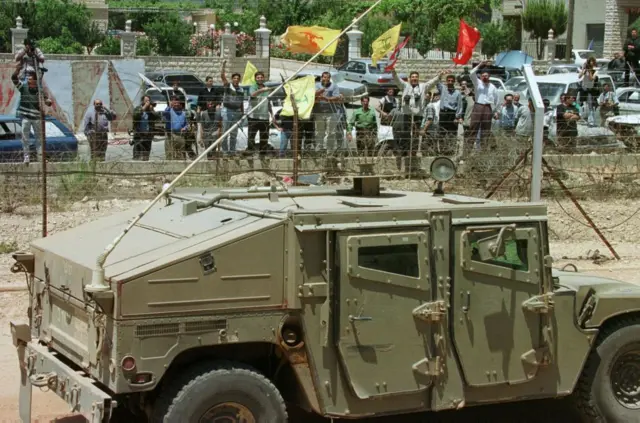 24 May 2000:  People wave Hezbollah and Lebanon flags on the Lebanese side of the border, in front of Israeli vehicles on the israeli side