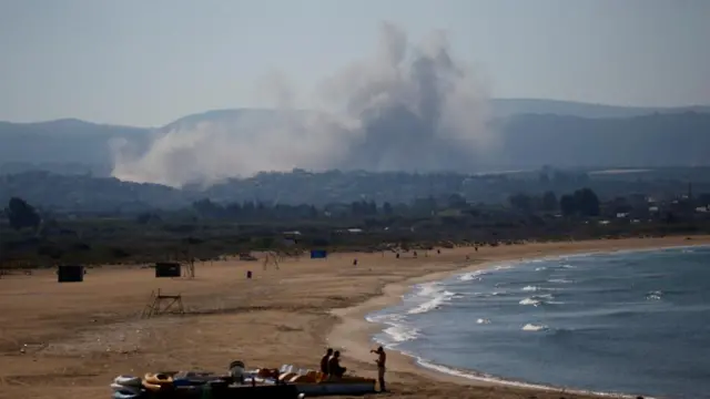 People spend time at a beach as smoke billows in the background over southern Lebanon